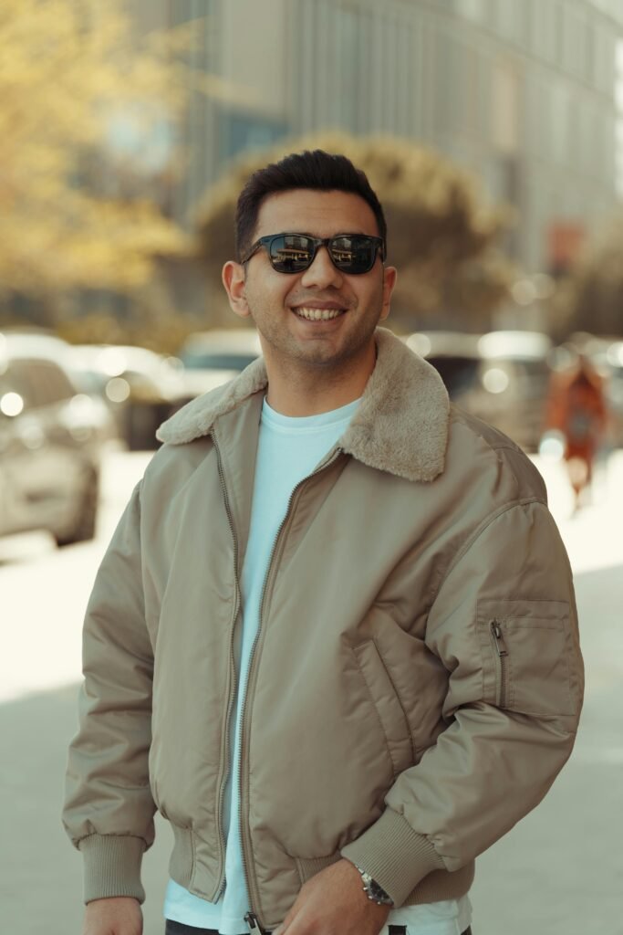 A man wearing sunglasses and outerwear smiles confidently on a sunny city street.